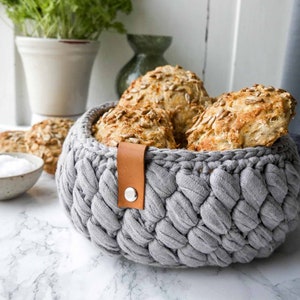 May include: A gray crocheted basket with a brown leather handle and a metal button. The basket is filled with four round bread rolls topped with sunflower seeds.
