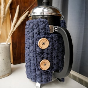 Pode incluir: Uma cafeteira de prensa francesa com uma capa de crochê azul marinho. A capa tem dois botões grandes de madeira. A cafeteira tem uma alça preta e uma tampa e base prateadas.