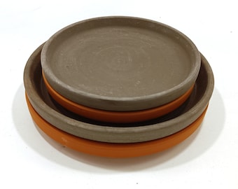 May include: Three terracotta plant saucers, one stacked on top of the other. The top saucer is a light brown colour, the middle saucer is a darker brown colour, and the bottom saucer is a bright orange colour.