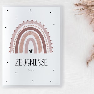 Könnte beinhalten: Ein weißer rechteckiger Druck mit einem braunen und rosa Regenbogen-Design und einem schwarzen Herzen. Das deutsche Wort "ZEUGNISSE" ist in Schwarz gedruckt, darunter der Name "Mira". Schwarze Punkte sind um die Ränder des Drucks verstreut.