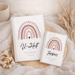 Könnte beinhalten: Zwei weiße Baby-Aufnahmehefte mit einem Regenbogen-Design in Brauntönen und einem schwarzen Herz. Das größere Buch trägt die Aufschrift "U-Heft" und das kleinere "Impfpass". Beide Bücher tragen den Namen "Mira Muster" und das Datum "01.08.2020".