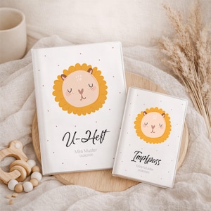 Könnte beinhalten: Zwei weiße Baby-Gesundheitshefte mit einer Löwenillustration. Das größere Buch trägt die Aufschrift "U-Heft" und das kleinere "Impfpass". Beide Bücher tragen den Namen "Mira Muster" und das Datum "01.08.2020".