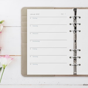Könnte beinhalten: Ein beiger Planer mit einer weißen Seite, die für Januar 2021 geöffnet ist. Die Seite hat ein wöchentliches Kalenderlayout mit den Wochentagen in deutscher Sprache. Der Planer hat eine silberne Ringbindung.