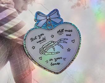 May include: A holographic heart-shaped sticker with a blue bow and a skeleton hand reaching out. The sticker says "But you would still miss me in your bones."
