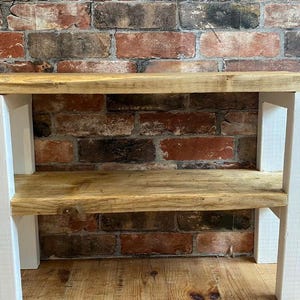 May include: A two-tiered shelf unit made of reclaimed wood with a white painted frame. The shelf unit is standing on a wooden floor in front of a brick wall.