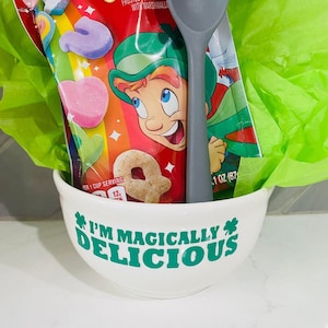 May include: A white ceramic bowl with the text "I'M MAGICALLY DELICIOUS" in green, filled with a bag of Lucky Charms cereal, a gray spoon, and green tissue paper. The cereal bag features a leprechaun and colorful marshmallows.