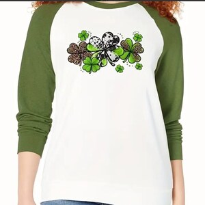 May include: A white long-sleeved shirt with green and black shamrock designs on the front. The shirt has a green and white color block design with green sleeves.