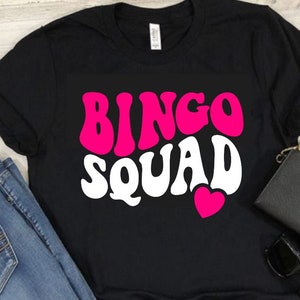 May include: Black t-shirt with a pink and white graphic that says "Bingo Squad" with a pink heart.