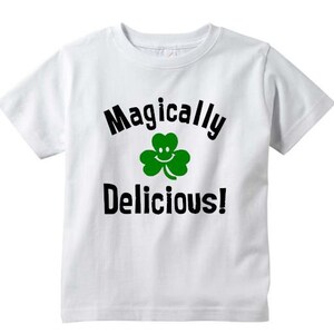 May include: White t-shirt with black text that reads "Magically Delicious!" and a green shamrock with a smiley face.