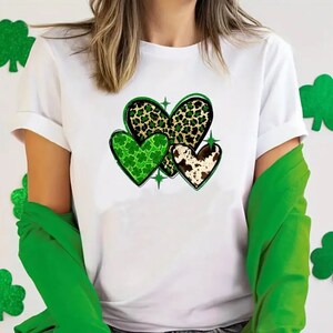 May include: White t-shirt with a design of three hearts. The hearts are patterned with green shamrocks, leopard print, and cowhide.