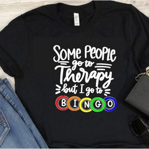 May include: Black t-shirt with white text that reads "Some people go to therapy but I go to BINGO" with colorful bingo balls.