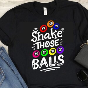 May include: Black t-shirt with white text that says "Shake Those Balls" and colorful bingo balls with numbers printed on them.