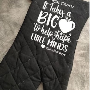 May include: Black quilted oven mitt with white text that reads "It Takes a BIG to help shape LITTLE MINDS". The text also includes "Miss Christy" and "The Wiley Boys".