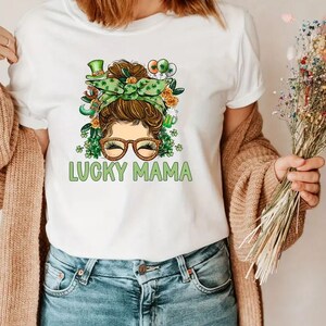 May include: White t-shirt with a green, brown, and gold illustration of a woman with a messy bun, glasses, and a shamrock headband. The text "LUCKY MAMA" is printed below the illustration.