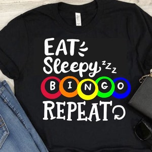 May include: Black t-shirt with white text that reads "EAT, SLEEPY, BINGO, REPEAT". The text is surrounded by five colorful circles with the letters "B", "I", "N", "G", and "O" in white.