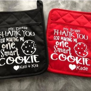 May include: Two black and red oven mitts with white text that reads "Thank you for making me one smart cookie". The black mitt has the text "Ms. Dirkse" and "Kalli & Kya" and the red mitt has the text "Ms. Briggs" and "Kade".