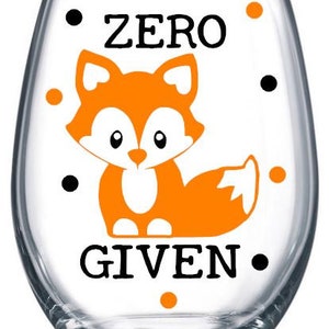 May include: A clear glass wine glass with a white background and black polka dots. The glass has a cartoon orange fox with black eyes and a white belly. The text "ZERO GIVEN" is printed in black on the glass.