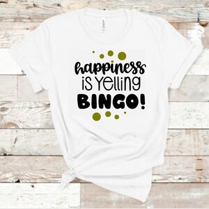May include: White t-shirt with the phrase "happiness is Yelling BINGO!" in black lettering and green dot accents. The shirt is made of a soft material and is knotted at the bottom.