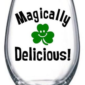 May include: A clear glass wine glass with a black and green design that says "Magically Delicious!" with a green shamrock with a smiley face.