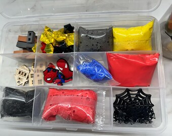 May include: A clear plastic storage box with multiple compartments filled with various craft supplies. The compartments contain red, blue, black, and yellow slime, black spider webs, red and blue glitter, and small wooden houses and figures.