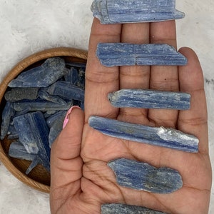 May include: A collection of blue kyanite crystal points, some are long and thin, others are shorter and wider. The crystals are arranged in a hand, with a bowl of smaller kyanite crystals in the background.
