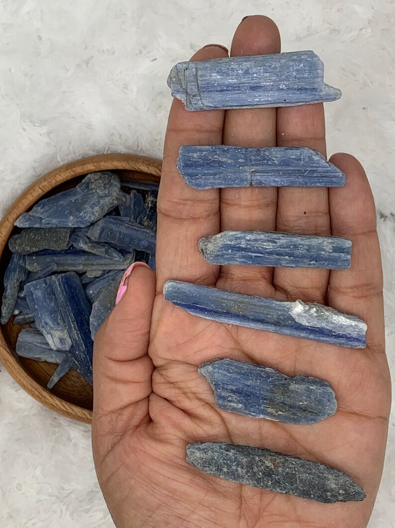 May include: A collection of blue kyanite crystal points, some are long and thin, others are shorter and wider. The crystals are arranged in a hand, with a bowl of smaller kyanite crystals in the background.