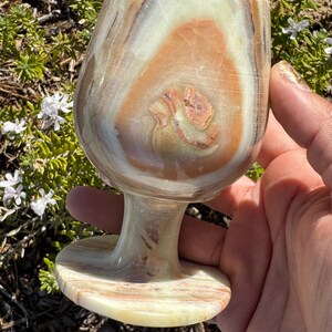 May include: A hand holds a large, intricately patterned, light green, brown, and white onyx goblet. The goblet has a wide, round bowl and a short, sturdy stem.