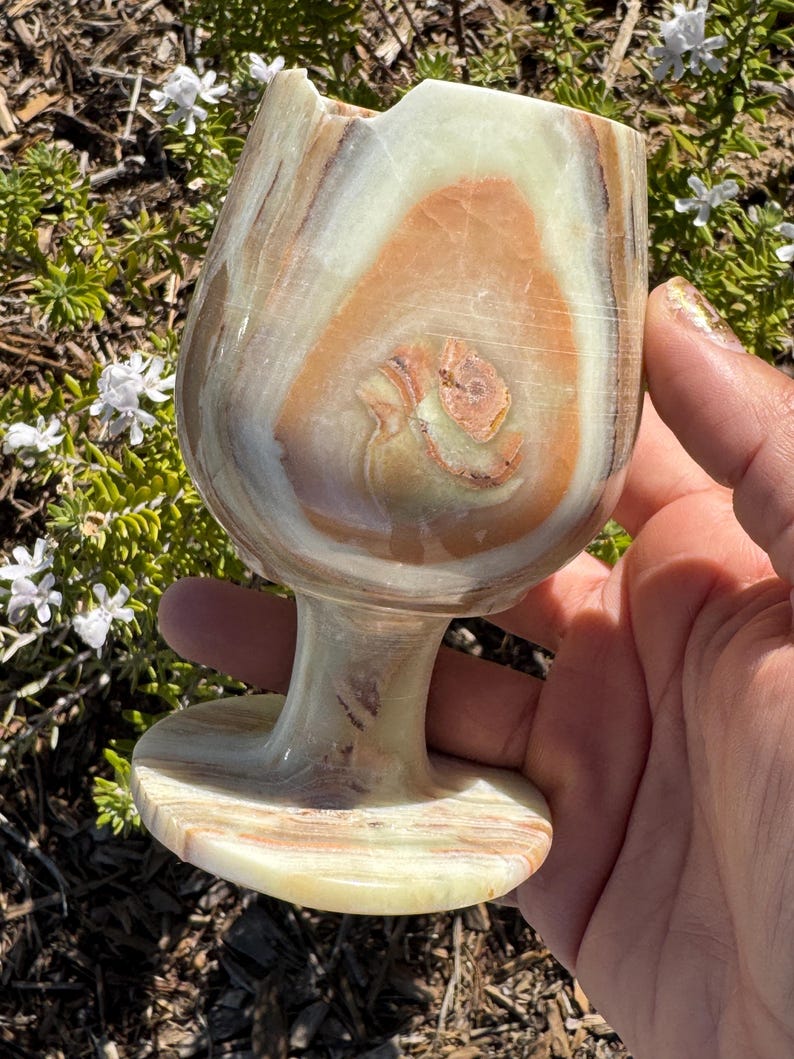 May include: A hand holds a large, intricately patterned, light green, brown, and white onyx goblet. The goblet has a wide, round bowl and a short, sturdy stem.