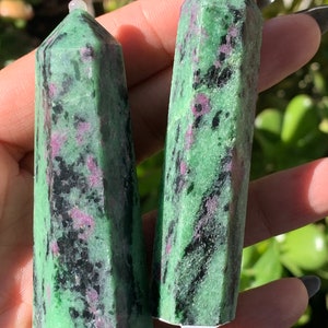 Könnte beinhalten: Zwei grüne und schwarze Rubin-Zoisit-Kristalltürme. Die Kristalle haben eine polierte, facettierte Form mit einer spitzen Spitze. Die grüne Farbe ist mit schwarzen und rosaroten Einschlüssen durchsetzt. Die Kristalle werden in einer Hand gehalten.