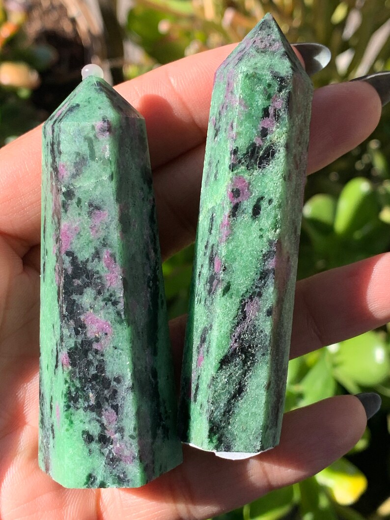 Peut inclure: Deux tours de cristal de rubis zoisite vertes et noires. Les cristaux ont une forme polie et facett&eacute;e avec un sommet pointu. La couleur verte est entrecoup&eacute;e d'inclusions noires et rouge-ros&eacute;. Les cristaux sont tenus dans une main.