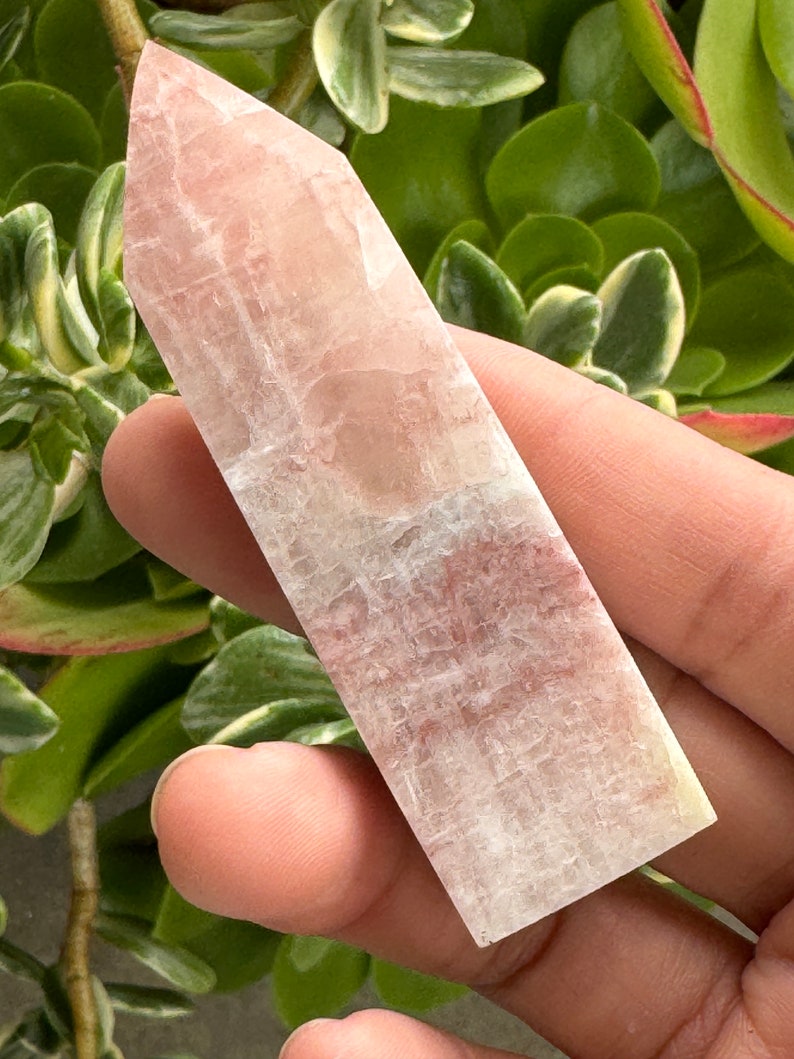 May include: A clear, white, and pink crystal point with a smooth, polished surface. The crystal is shaped like a long, thin pyramid.