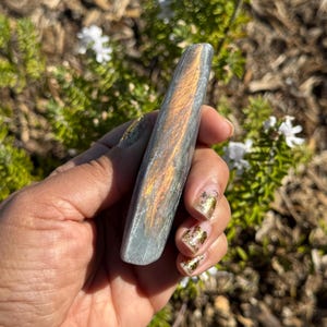 May include: A long, smooth, grey and iridescent crystal with a golden sheen. The crystal is shaped like a rounded rectangle and is held in a hand.