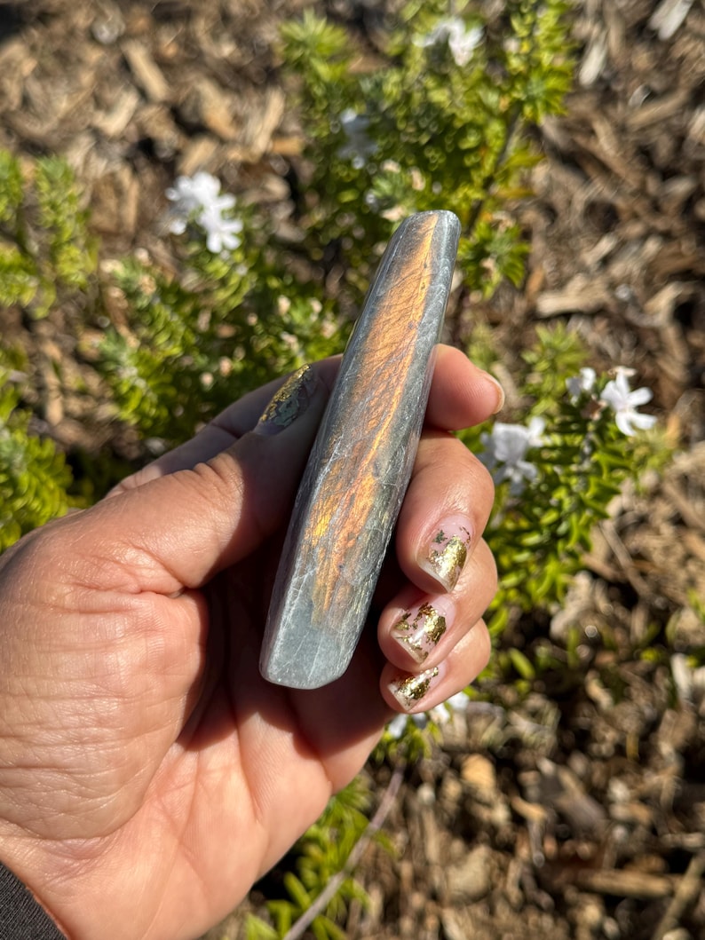 May include: A long, smooth, grey and iridescent crystal with a golden sheen. The crystal is shaped like a rounded rectangle and is held in a hand.