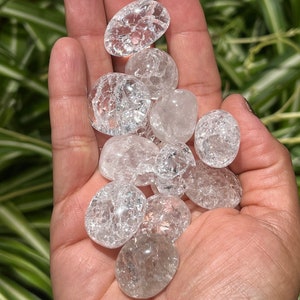 May include: A collection of clear quartz crystal tumbled stones with a cloudy, crackled appearance. The stones are smooth and polished, and they are held in a hand.