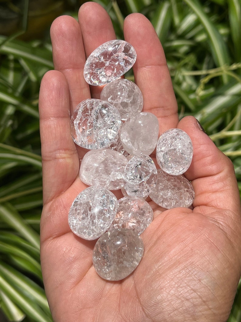 May include: A collection of clear quartz crystal tumbled stones with a cloudy, crackled appearance. The stones are smooth and polished, and they are held in a hand.