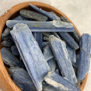 May include: A wooden bowl filled with blue kyanite crystal points. The crystals are long and slender, with a light blue color and a slightly rough texture.