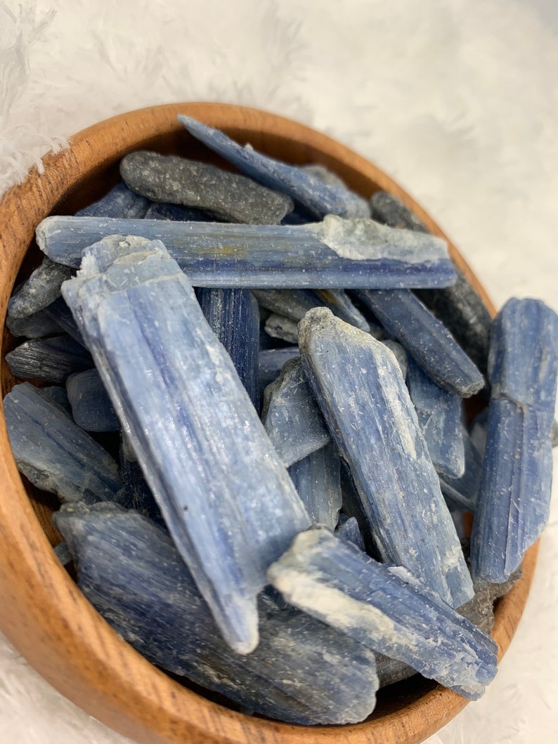May include: A wooden bowl filled with blue kyanite crystal points. The crystals are long and slender, with a light blue color and a slightly rough texture.