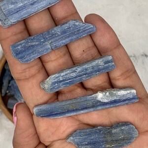May include: Five blue kyanite crystal points, each with a different shape and size, are displayed on a hand. The crystals are a light blue color and have a striated texture.
