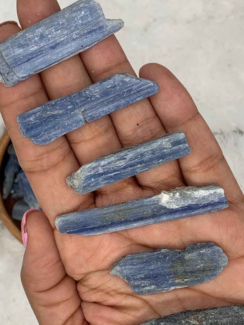 May include: Five blue kyanite crystal points, each with a different shape and size, are displayed on a hand. The crystals are a light blue color and have a striated texture.