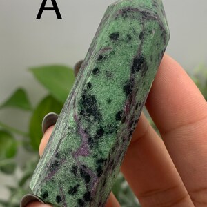 Peut inclure: Une pointe de cristal de zo&iuml;site verte et noire avec des veines violettes. Le cristal est tenu dans une main et a une forme polie et facett&eacute;e. La lettre "A" est visible dans le coin sup&eacute;rieur gauche. Le cristal est une pierre naturelle.
