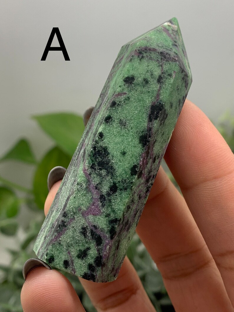 Peut inclure: Une pointe de cristal de zo&iuml;site verte et noire avec des veines violettes. Le cristal est tenu dans une main et a une forme polie et facett&eacute;e. La lettre "A" est visible dans le coin sup&eacute;rieur gauche. Le cristal est une pierre naturelle.