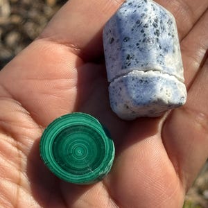 May include: A close-up of two polished stones. One is a round, green malachite stone with concentric rings. The other is a pointed, white and blue stone with black flecks.