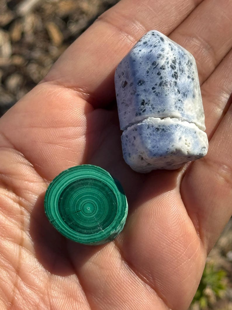 May include: A close-up of two polished stones. One is a round, green malachite stone with concentric rings. The other is a pointed, white and blue stone with black flecks.