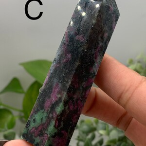 Peut inclure: Une tour de cristal rectangulaire fonc&eacute;e avec un sommet pointu. Le cristal pr&eacute;sente un m&eacute;lange de couleurs noires, rouge fonc&eacute; et vertes. La lettre "C" est visible dans le coin sup&eacute;rieur gauche. Le cristal est tenu dans une main.