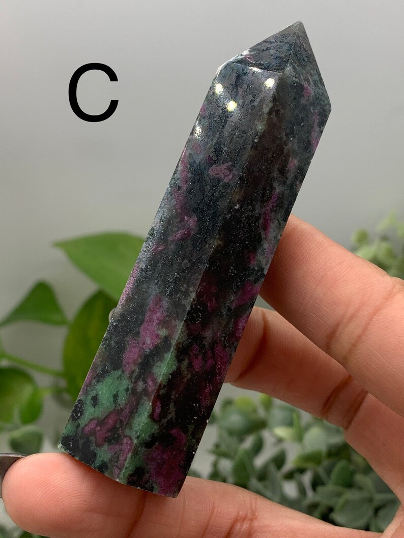 Peut inclure: Une tour de cristal rectangulaire fonc&eacute;e avec un sommet pointu. Le cristal pr&eacute;sente un m&eacute;lange de couleurs noires, rouge fonc&eacute; et vertes. La lettre "C" est visible dans le coin sup&eacute;rieur gauche. Le cristal est tenu dans une main.