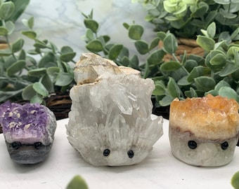 Puede incluir: Tres figuritas de erizo de cristal. La primera es de amatista púrpura, la segunda de cuarzo transparente y la tercera de citrino amarillo-naranja.