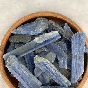 May include: A wooden bowl filled with blue kyanite crystal chips. The crystals are a light blue color with a white, striated pattern.