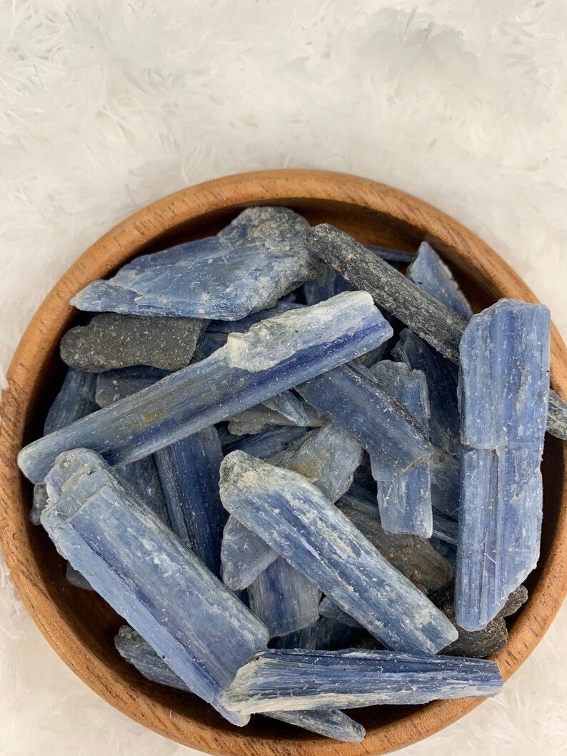 May include: A wooden bowl filled with blue kyanite crystal chips. The crystals are a light blue color with a white, striated pattern.
