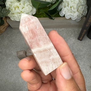 May include: A pale pink, translucent, rectangular crystal point with striations. The crystal is being held in a hand.