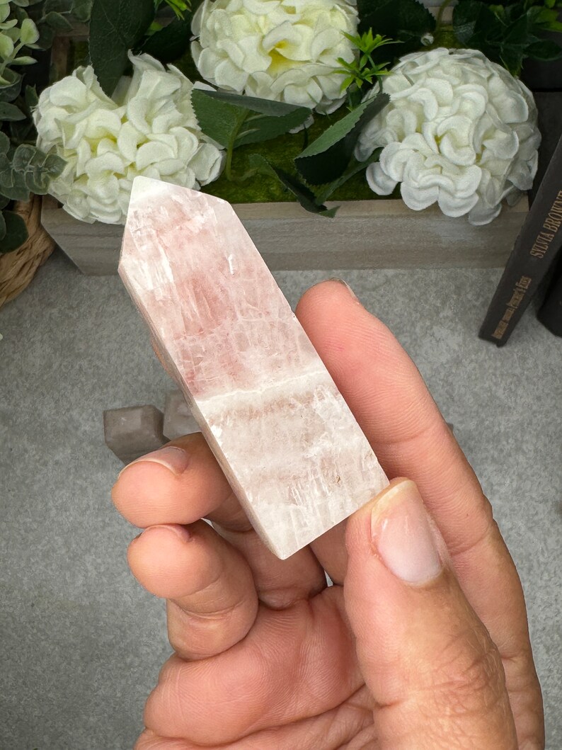 May include: A pale pink, translucent, rectangular crystal point with striations. The crystal is being held in a hand.